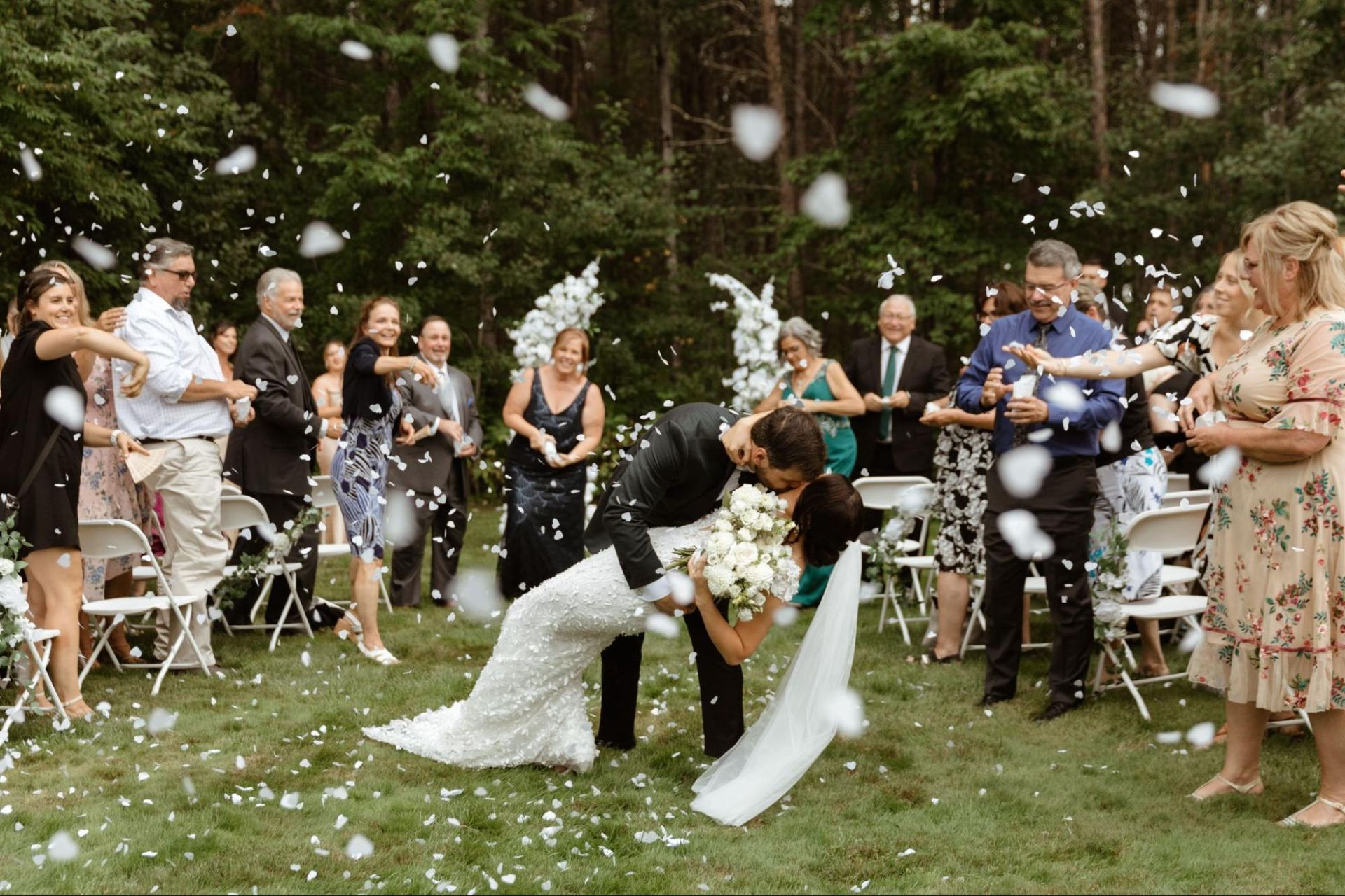 A bride and groom touch noses tenderly outdoors, both smiling, with a blurred water background.
