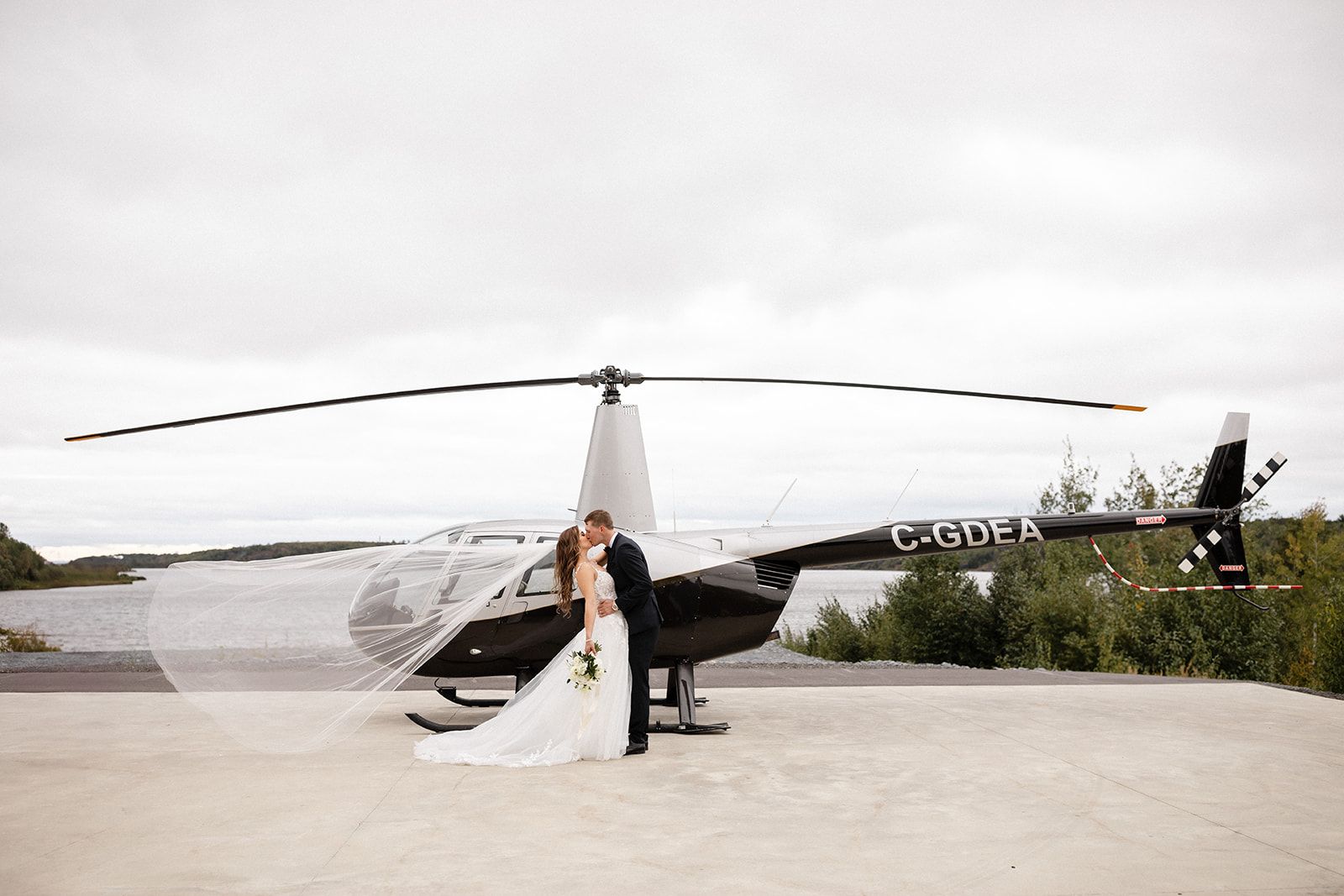 A couple in wedding attire embraces in front of a helicopter parked on a concrete pad by a body of water.