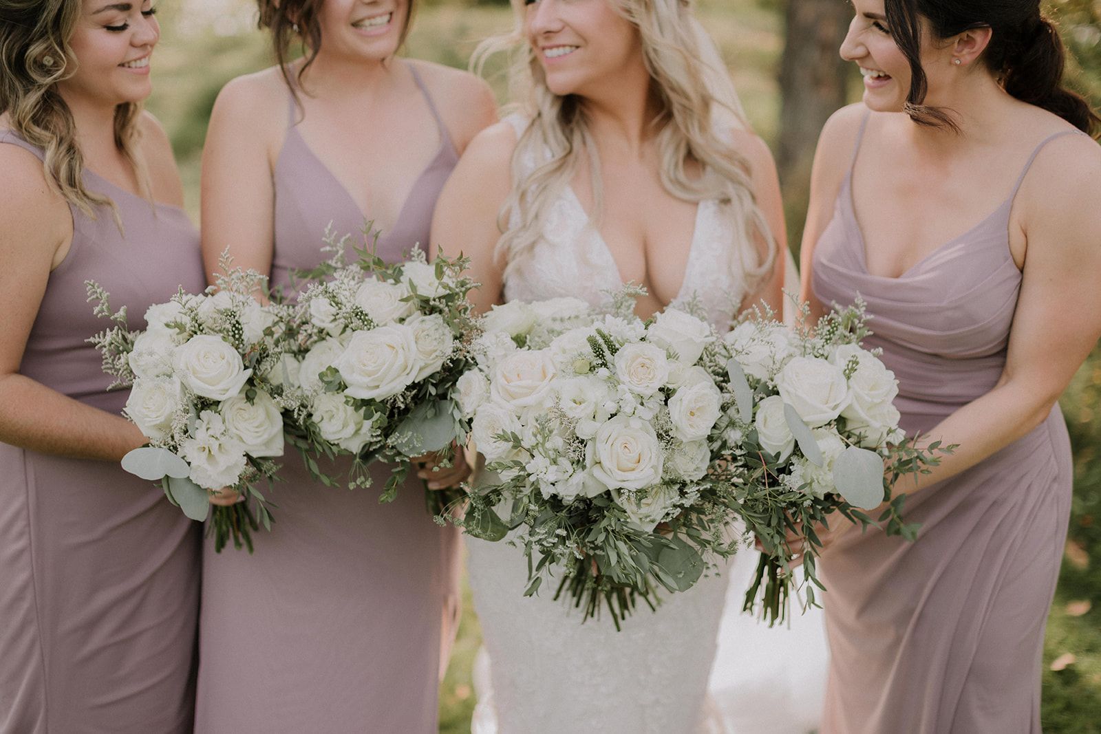 A bride and three bridesmaids in mauve dresses holding large white floral bouquets and smiling in an outdoor setting.