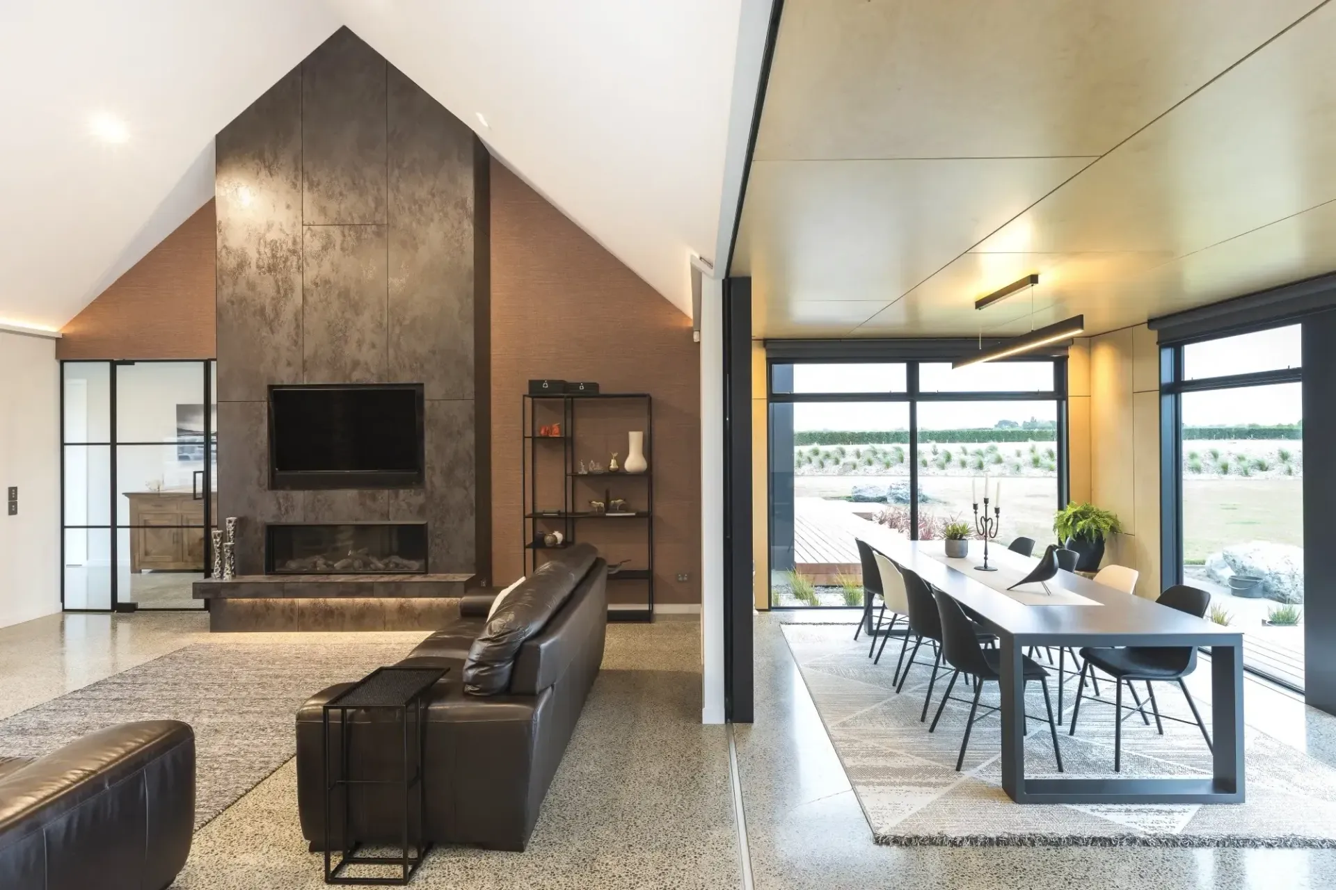 Modern living room with fireplace, TV, and dining area. Neutral tones, large windows, leather couch.