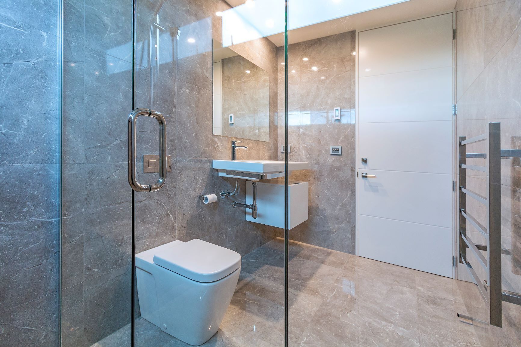 Modern bathroom with glass shower, gray tile, white toilet and sink, and a white door.