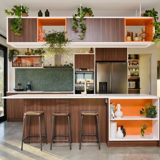 Modern kitchen with wood cabinets, orange accents, and hanging plants. Bar stools at island.