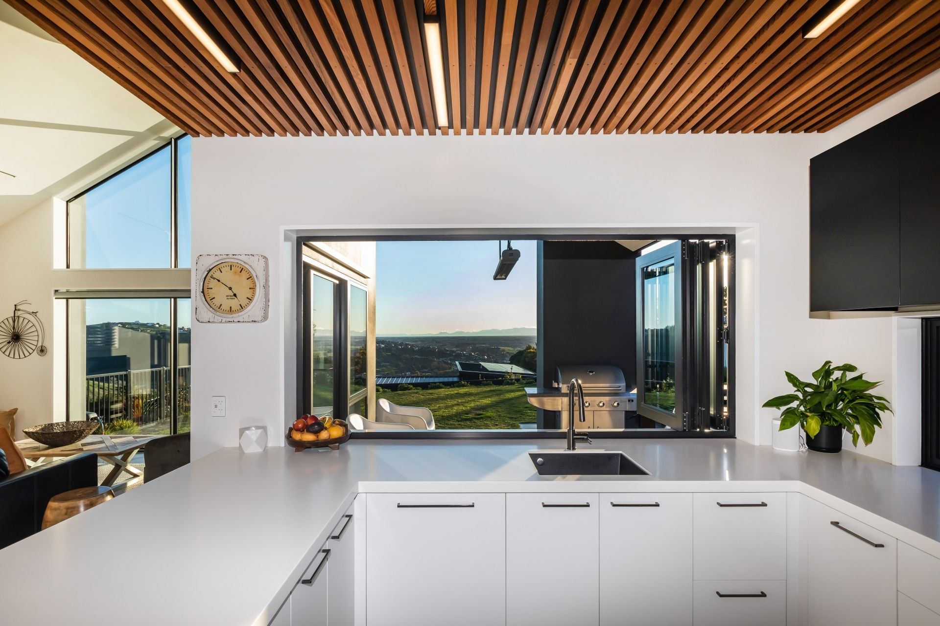 Modern kitchen with white cabinets, wooden ceiling, and large window opening to a grill and view.