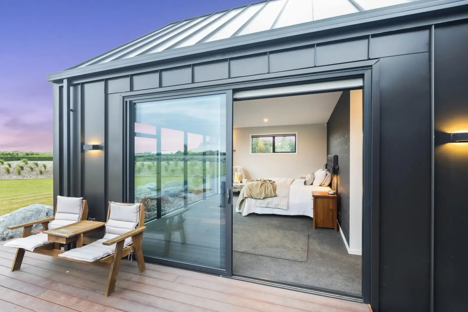 A modern black home with open sliding door to bedroom, outdoor chairs on deck, vineyard view.