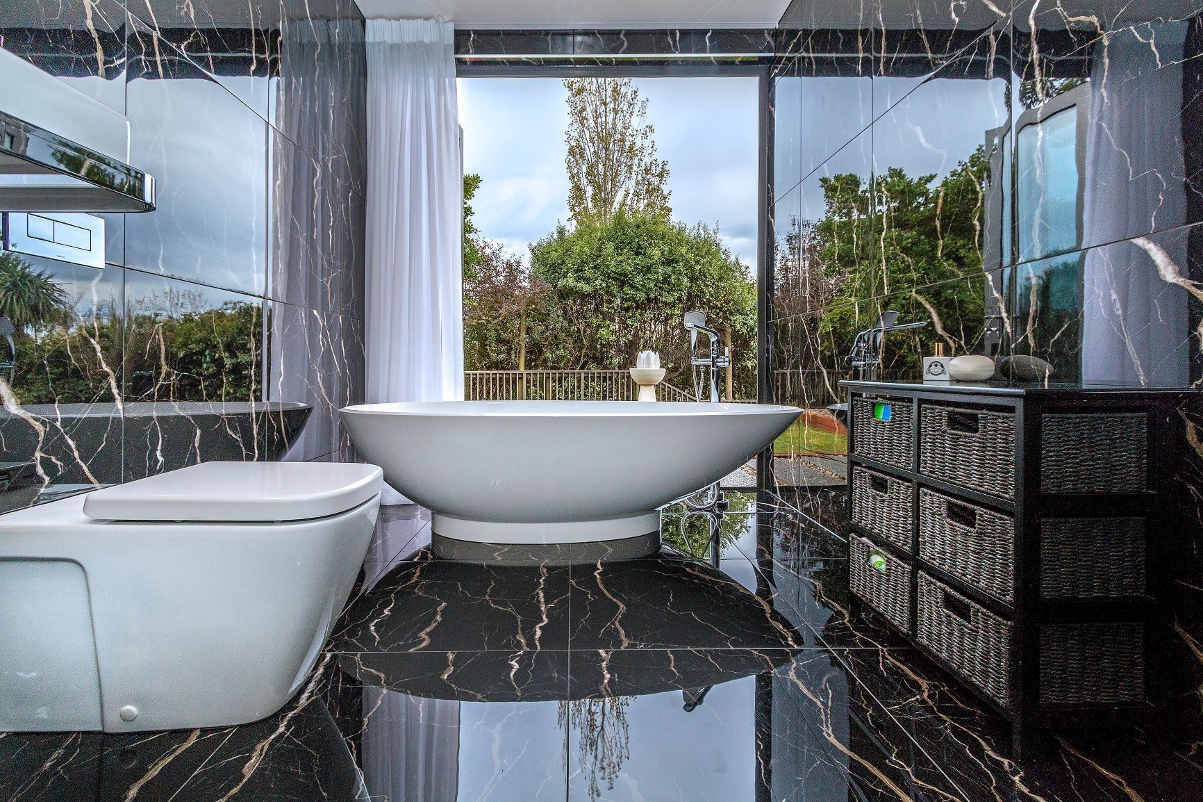 Modern bathroom with white tub, toilet, and black marble tile floor; large window with garden view.