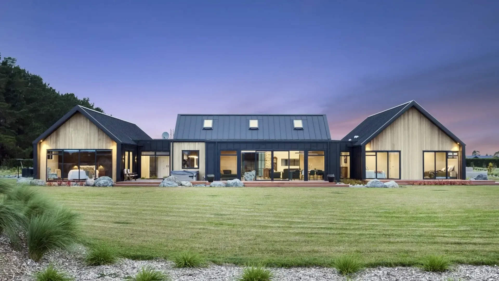 Modern single-story home with dark metal roof, light wood siding, and expansive windows on a green lawn.