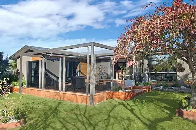 House with wooden deck and patio, surrounded by grass and a colorful tree, against a blue sky.