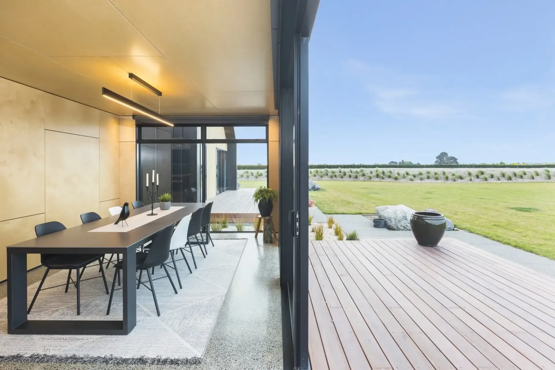 Dining room with long table, chairs, and open doors to a deck and green field under a blue sky.