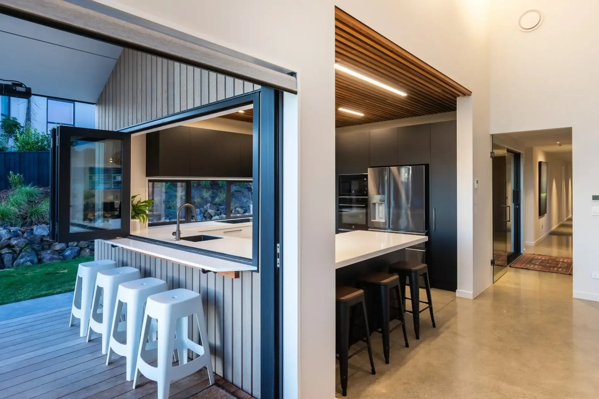 Modern kitchen with an outdoor bar; white stools, black countertops, and an open window.