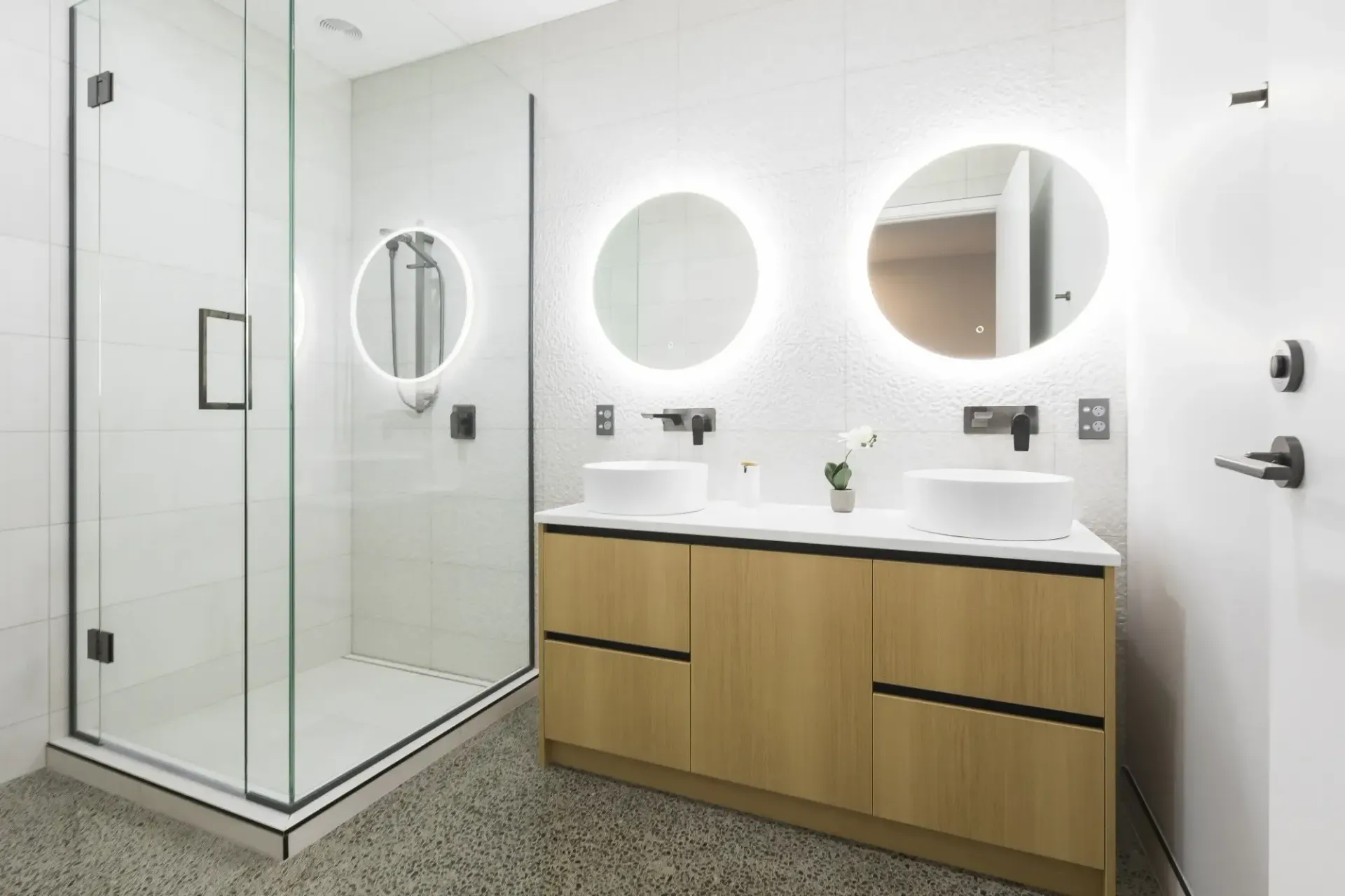 Modern bathroom with glass shower, vanity with two sinks, and round mirrors with lights.