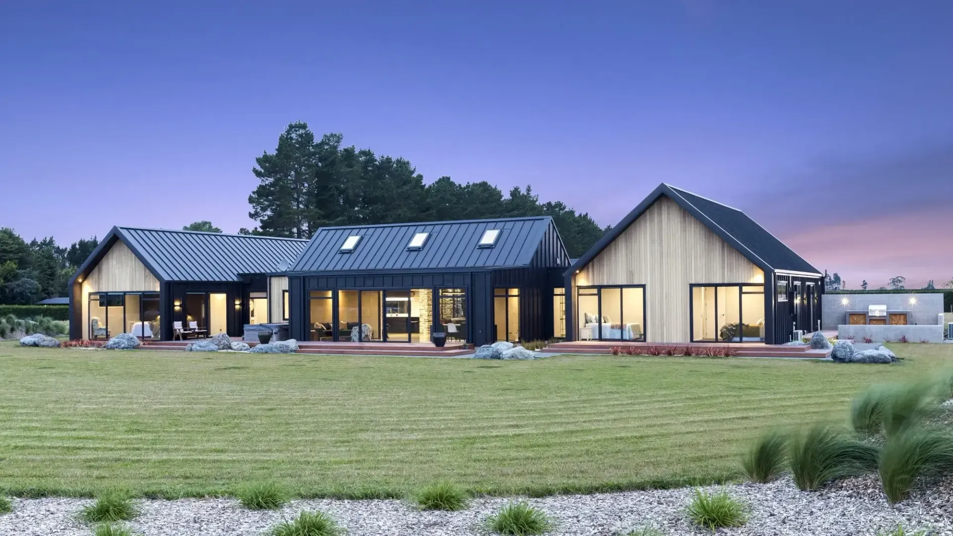Modern home with black siding, skylights, and large windows set on a grassy lawn at dusk.