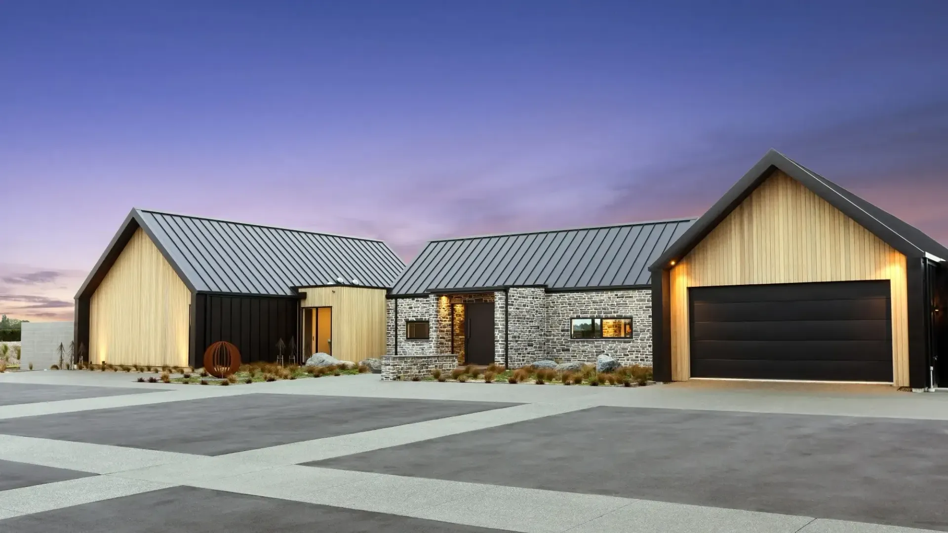 A modern home with natural wood and stone exterior, black trim, and a garage, under a dusk sky.