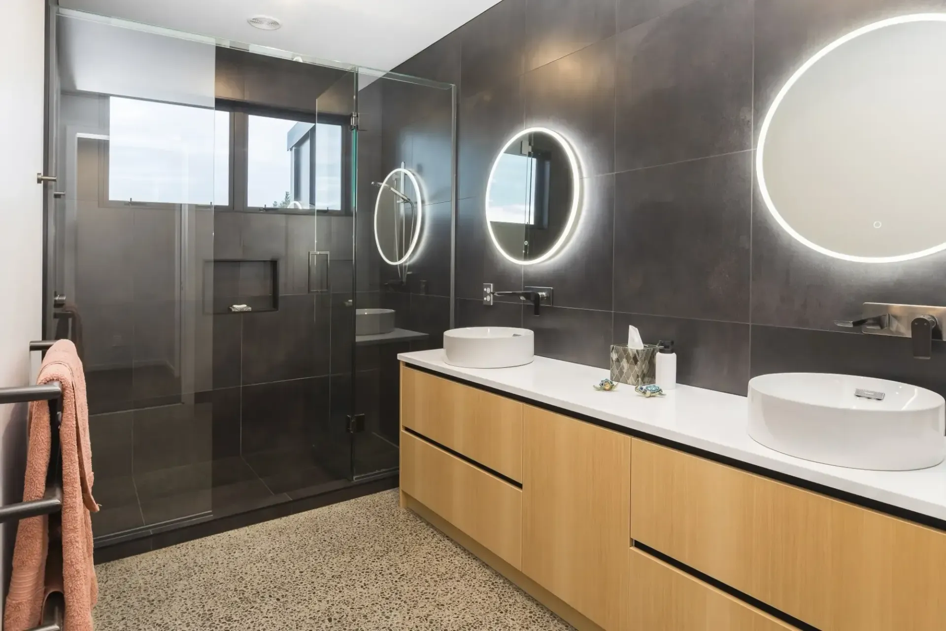 Modern bathroom with dark tile walls, wood vanity, round mirrors, and a glass shower.