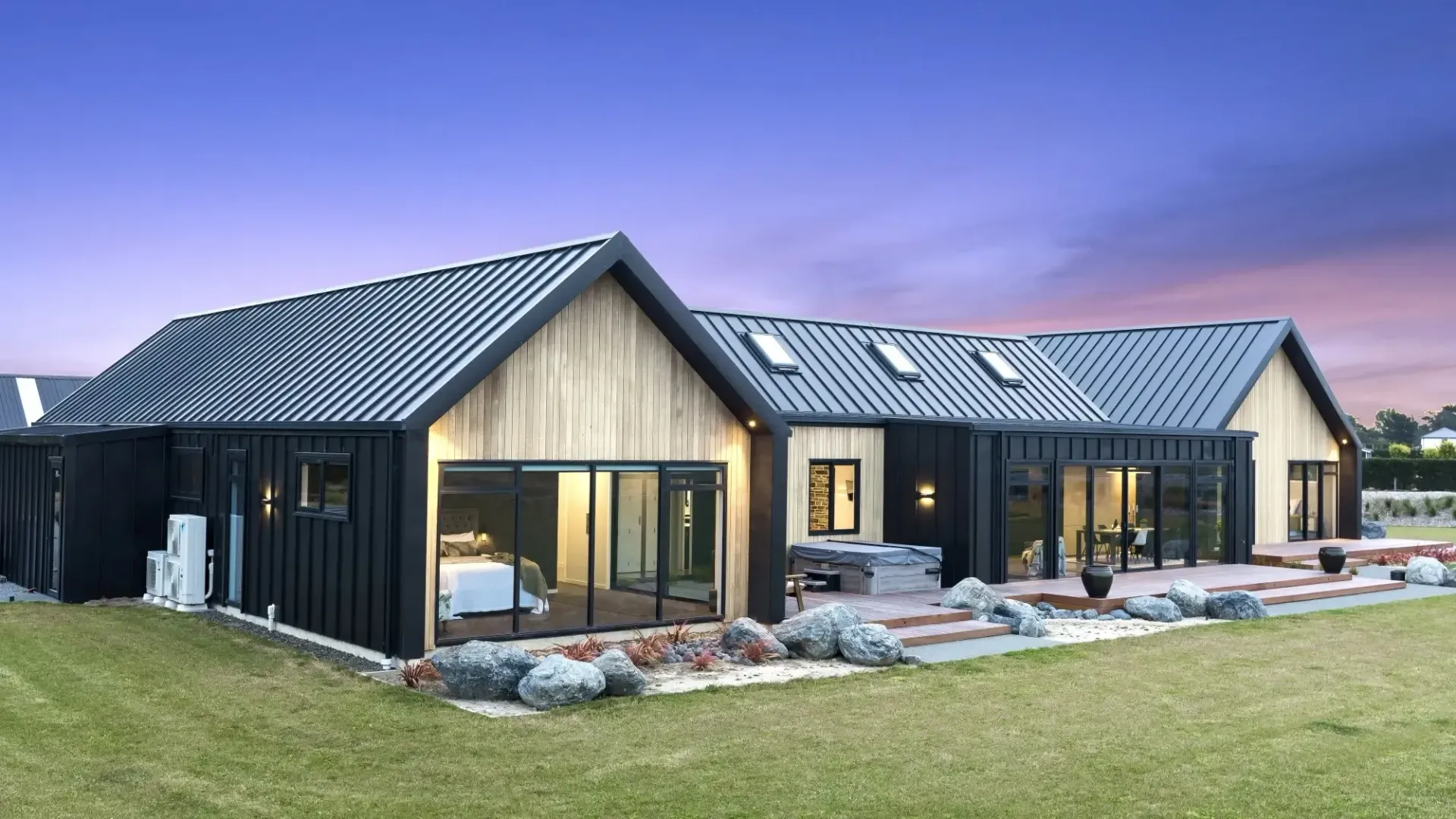 Modern black and wood-clad house with a dark roof, sliding doors, and a grassy yard at dusk.