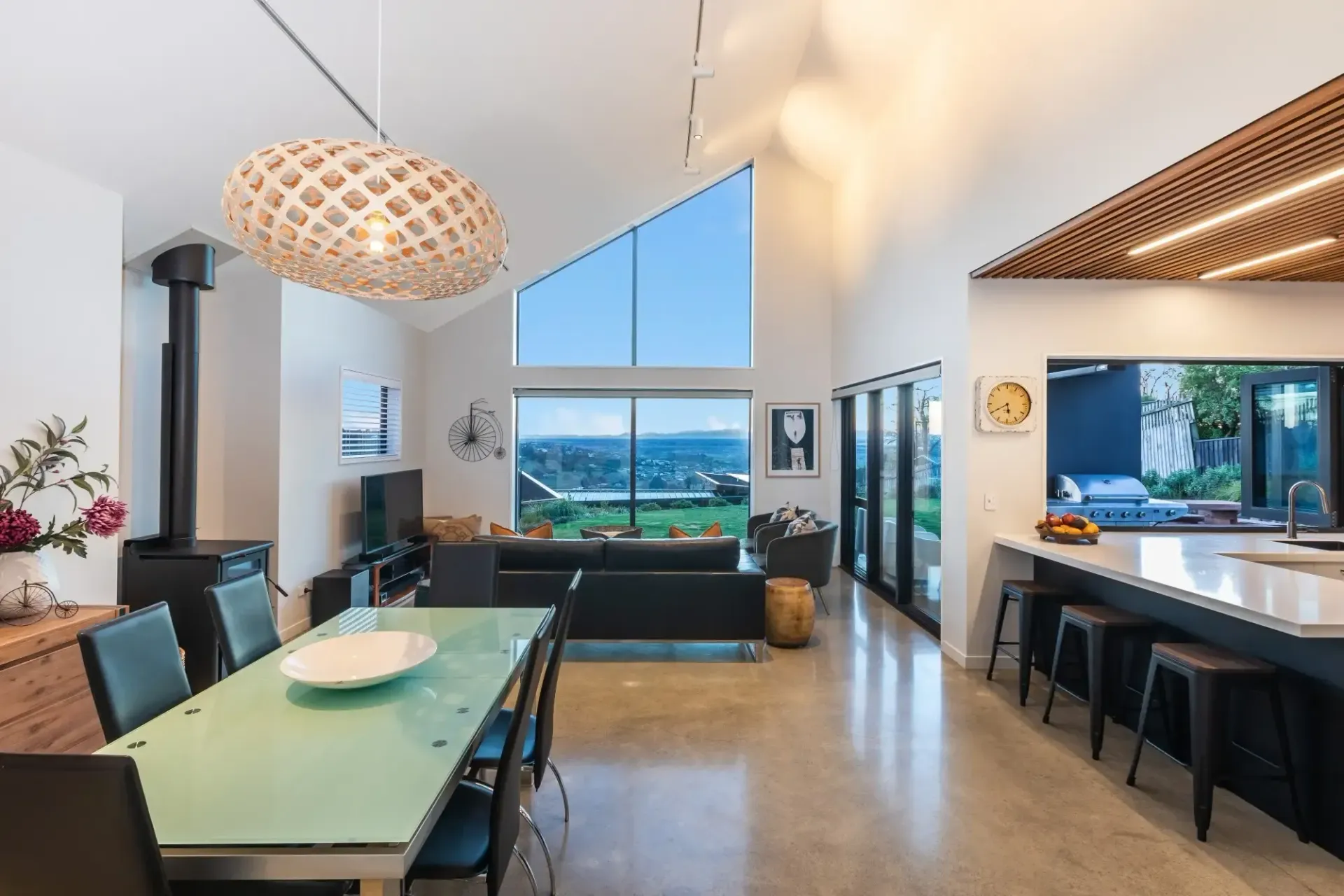 Modern living room with high ceilings, open to kitchen, featuring large windows and a dining table.
