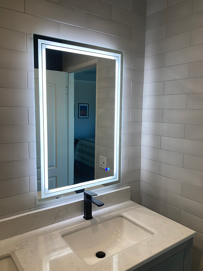 Lit rectangular bathroom mirror above a sink. The mirror reflects a bedroom and white door.
