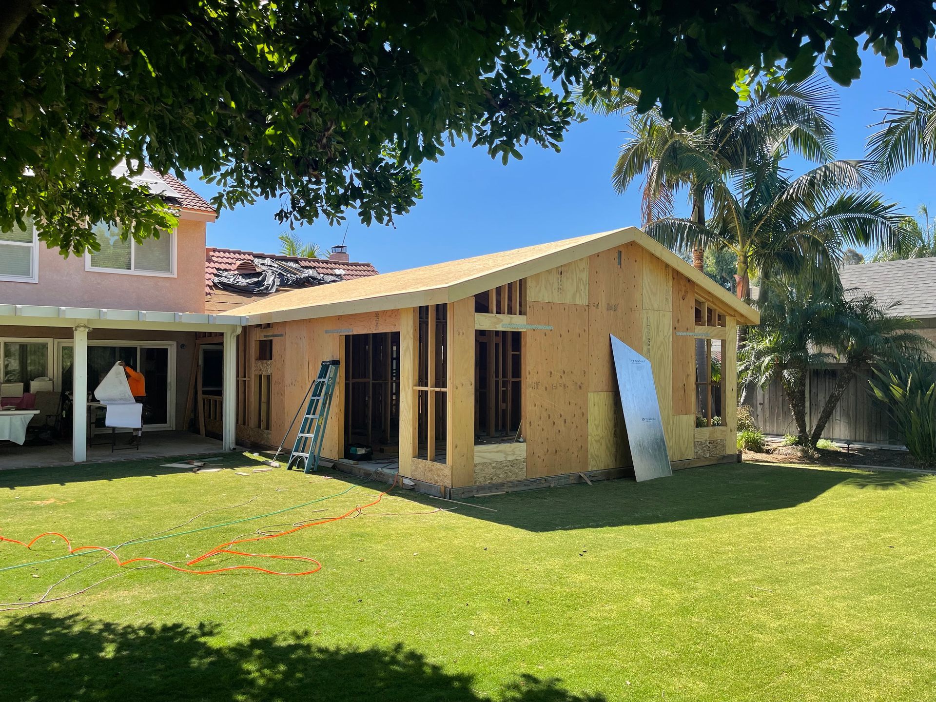 Construction of a new wood-framed room attached to a two-story house; work in progress on a sunny day.