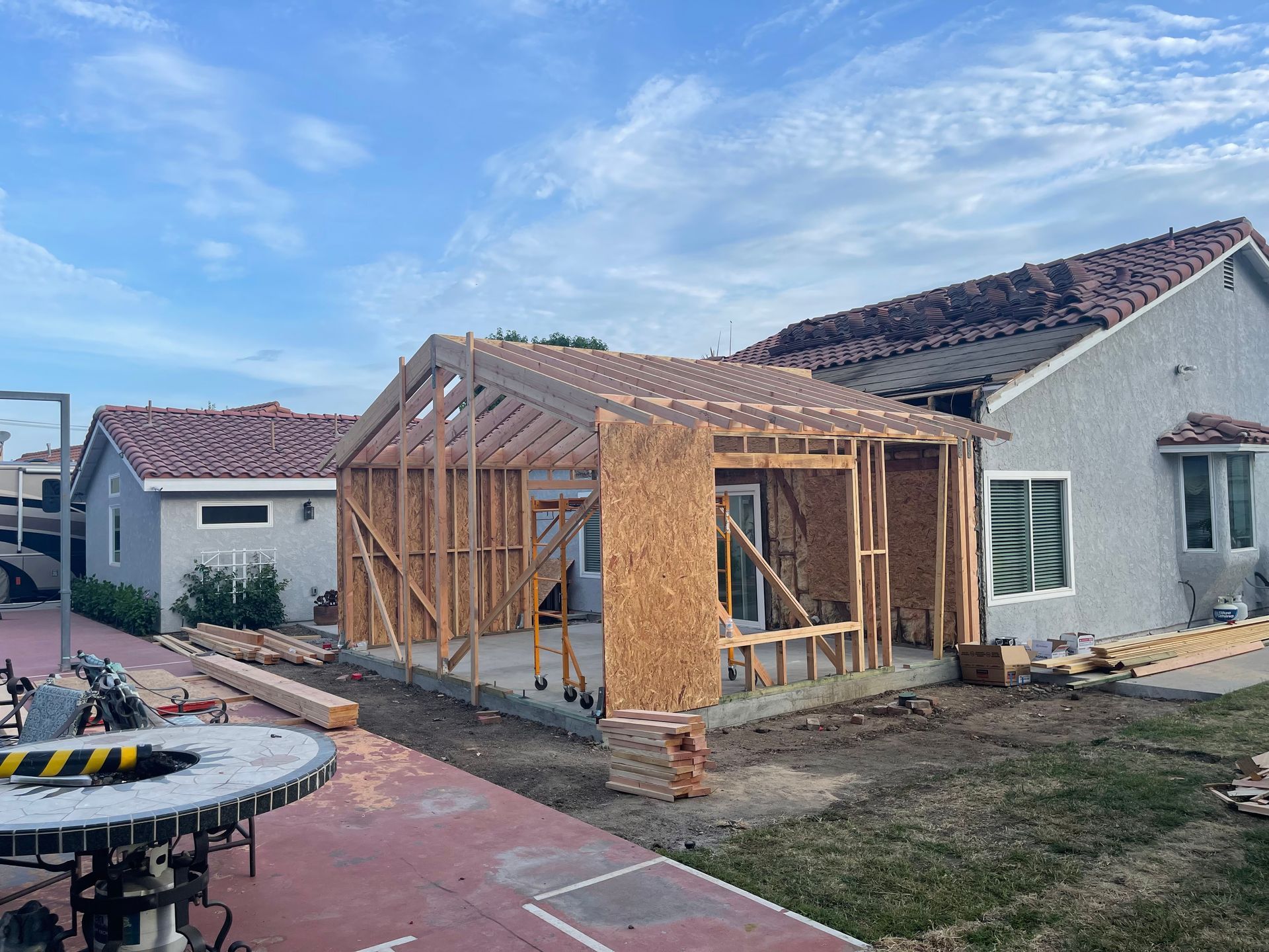 Construction in progress: Wooden frame extension attached to a house. Sky, grass, and red-tiled roofs visible.