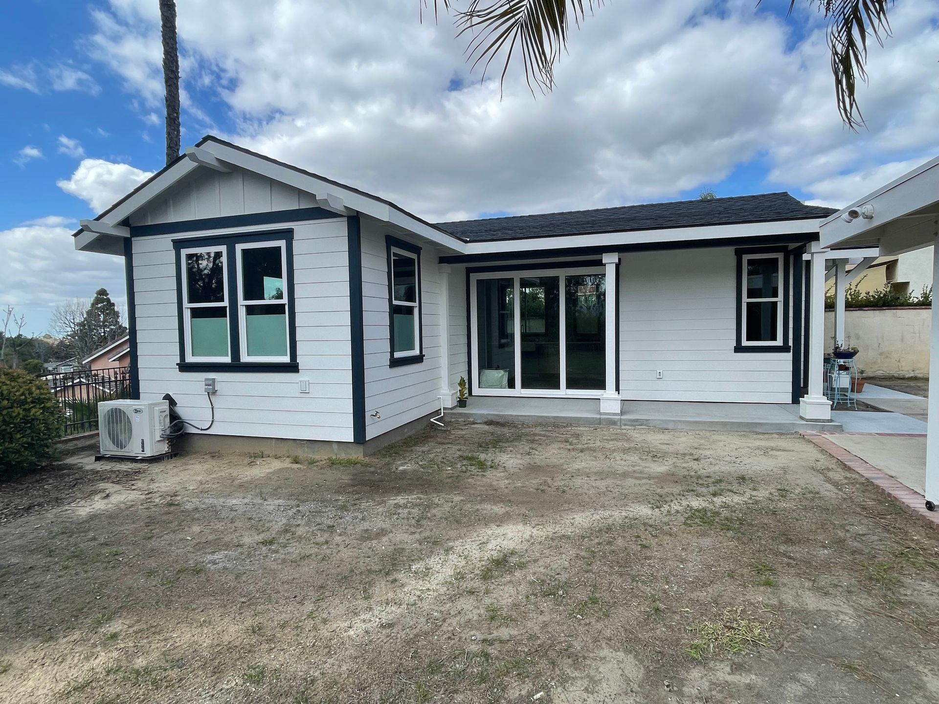 White house with dark trim, sliding glass doors, and an unkempt yard under a cloudy sky.