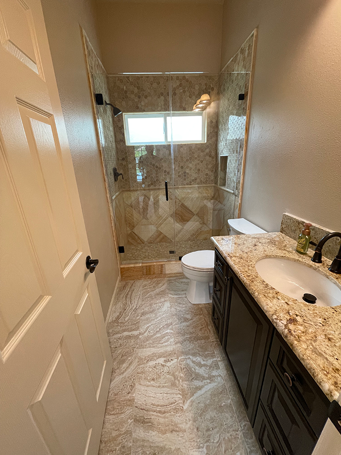 Bathroom with marble-patterned floor, brown vanity, toilet, and a glass shower with beige tile.