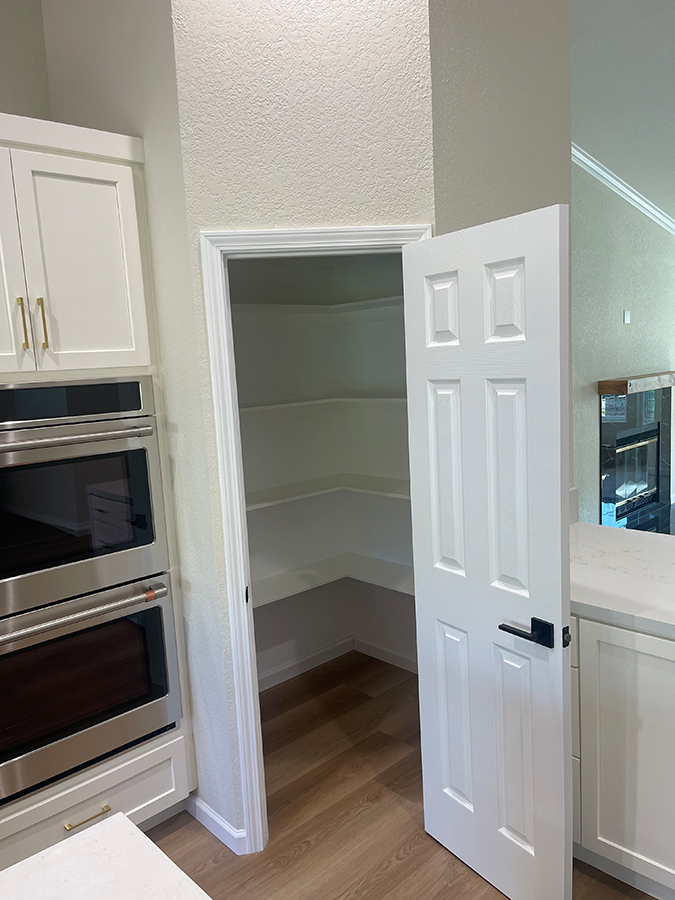 A white pantry door in a kitchen, open to reveal shelves. Adjacent to white cabinetry and appliances.