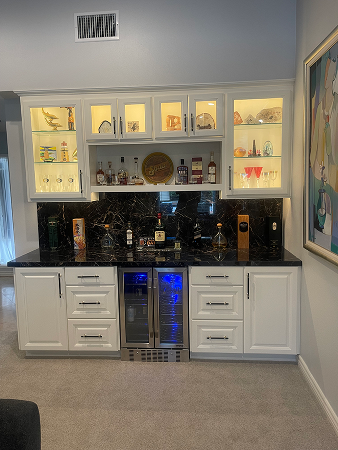 Home bar with black countertop, white cabinets, built-in refrigerator, and backlit shelves.