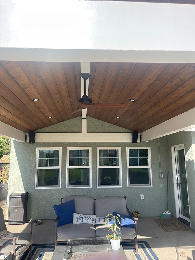 Outdoor patio with a wooden ceiling, ceiling fan, and gray couch with blue pillows.