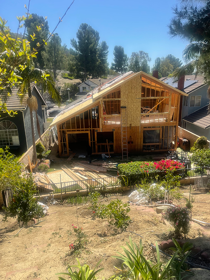 House under construction; wood frame visible. Workers, tools, and materials in a sunny, suburban yard.