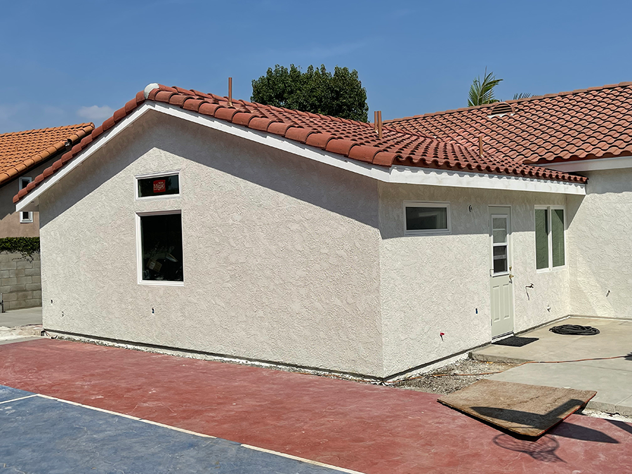 Tan stucco building with red tile roof, windows, and a door, set against a blue sky.