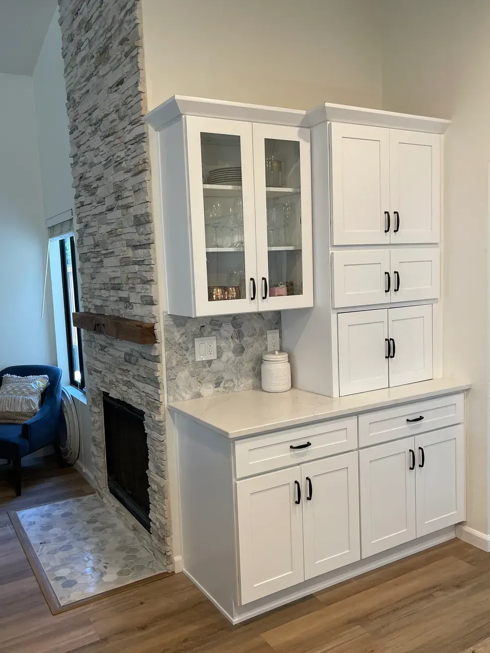 White kitchen cabinets next to a stone fireplace, with black hardware and a white countertop.