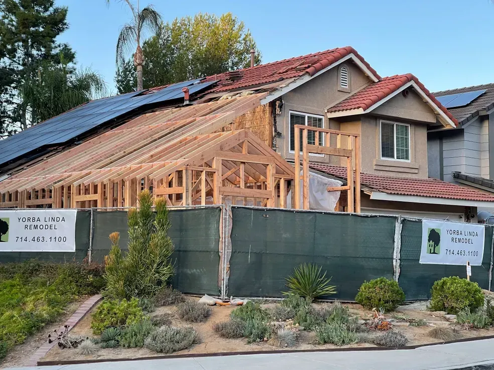 House under construction with exposed wooden framework, behind a green fence, solar panels on roof.