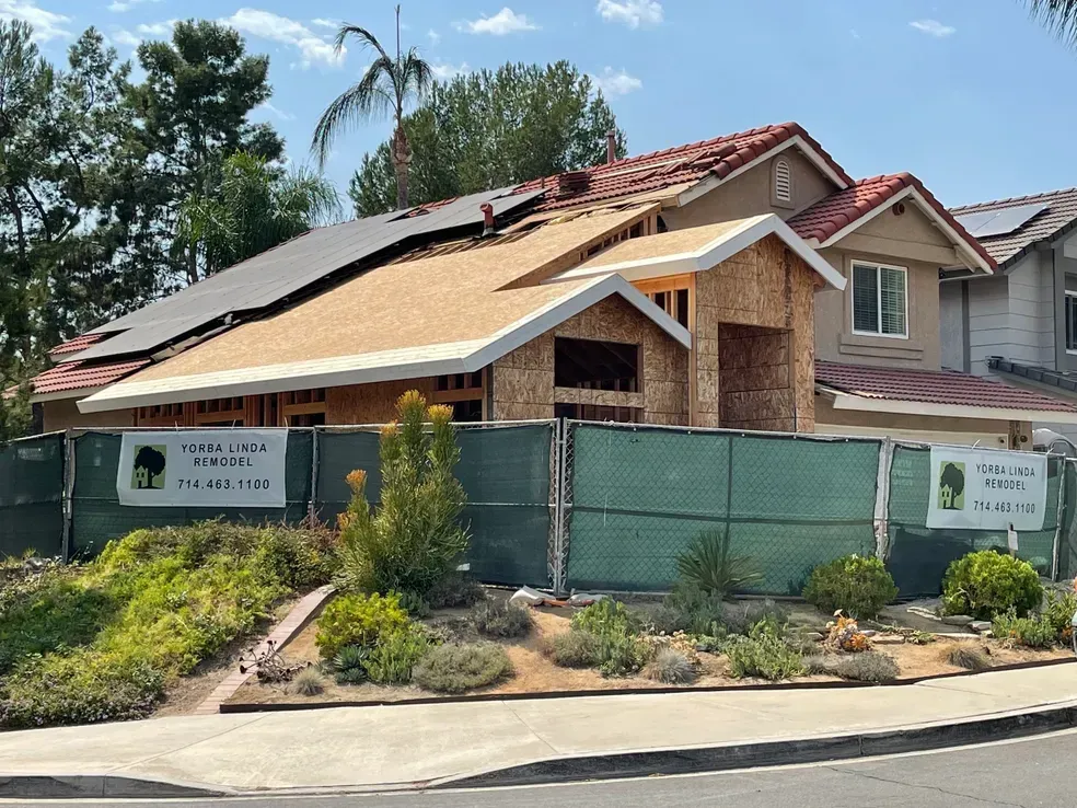 House under construction behind a green fence; unfinished roof, solar panels, and a sign.