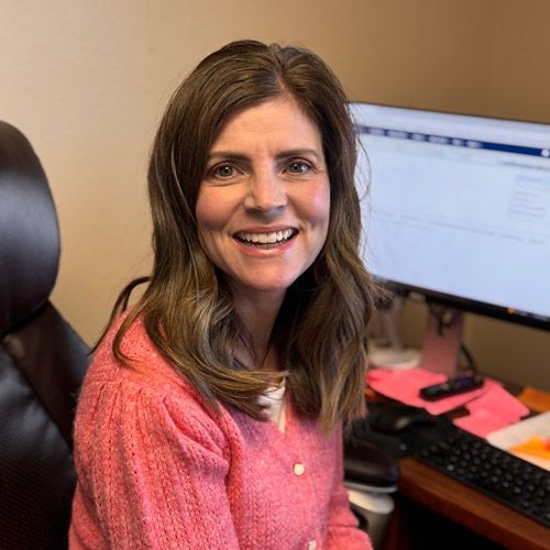 A person smiling while sitting at a desk in front of a computer monitor, wearing a pink knit sweater.