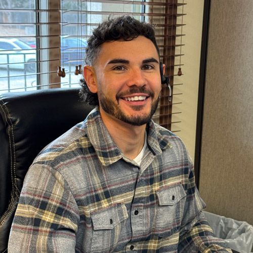 A person with a mullet hairstyle smiles while wearing a headset, seated at a desk in front of a window with blinds.