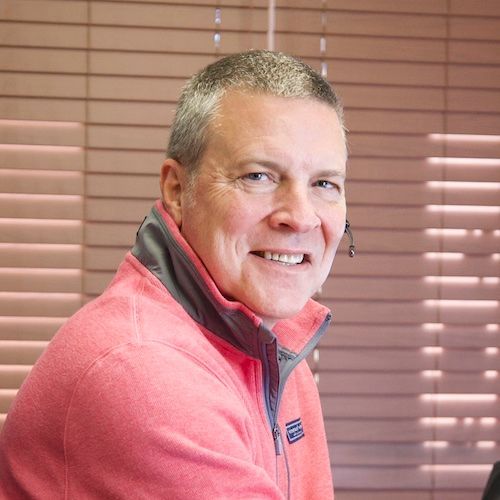 A person wearing a pink quarter-zip fleece and a headset smiles in front of closed horizontal window blinds.