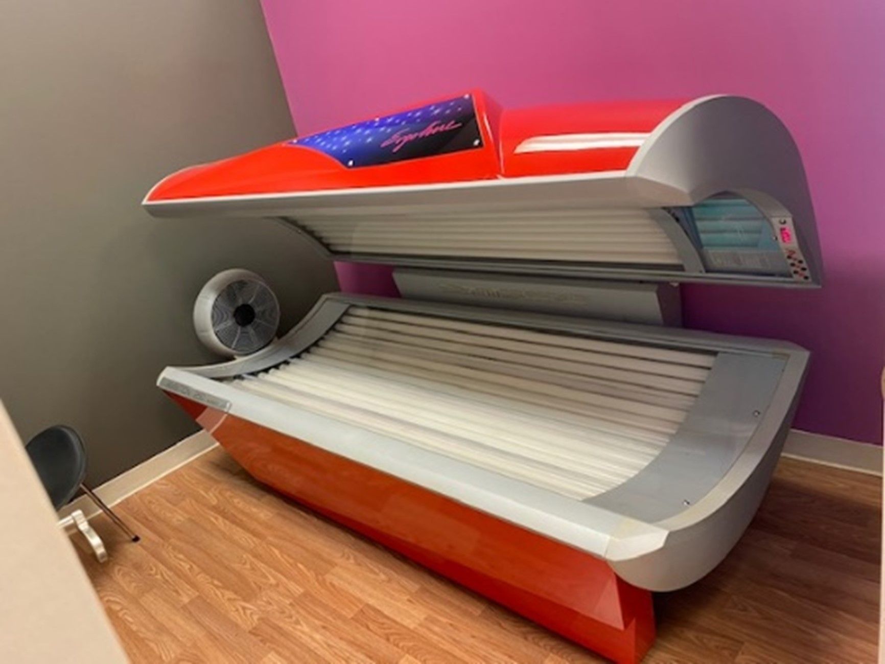 Red and gray tanning bed in a room with purple and pink walls, wooden floors, and a small fan.