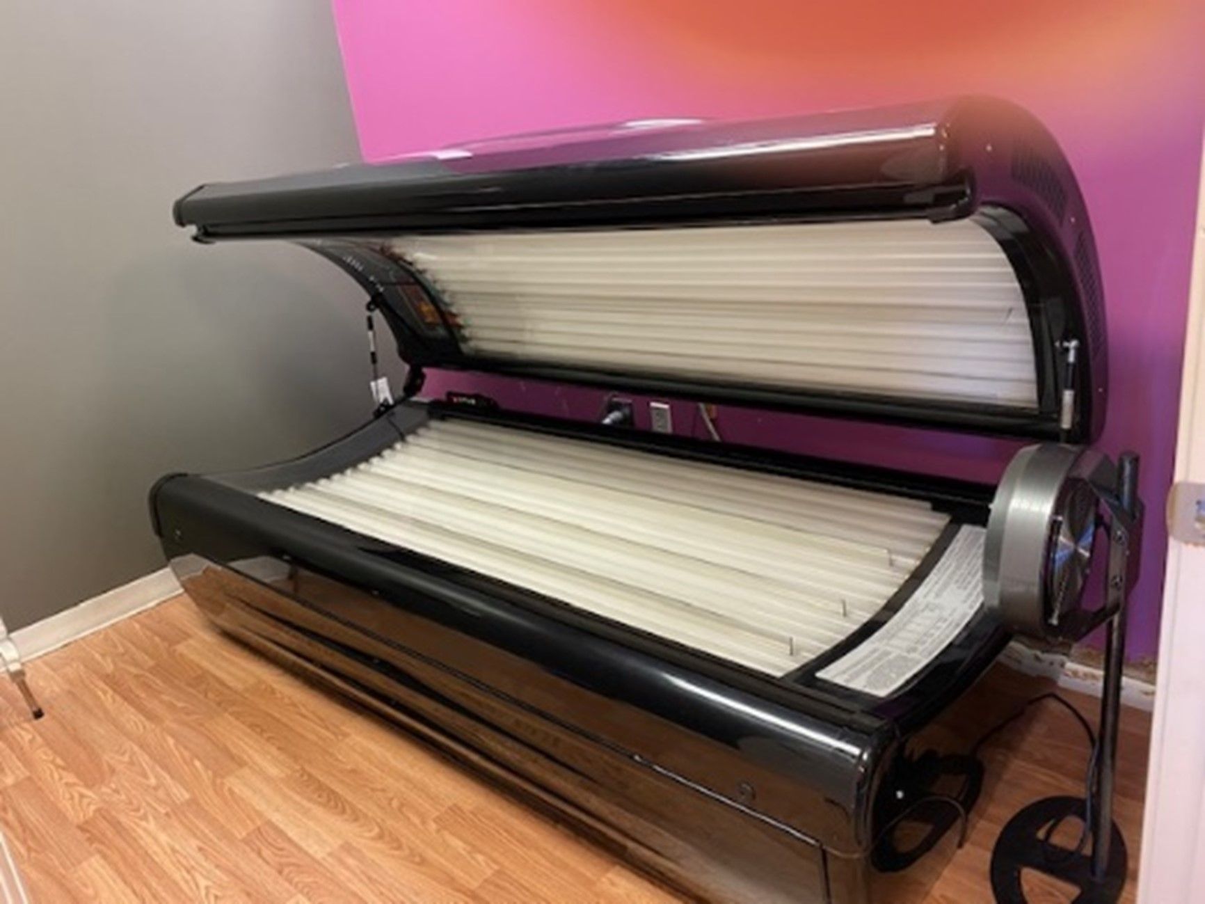 A black tanning bed with white fluorescent bulbs, set on a wooden floor in a salon. The bed is open and ready for use.