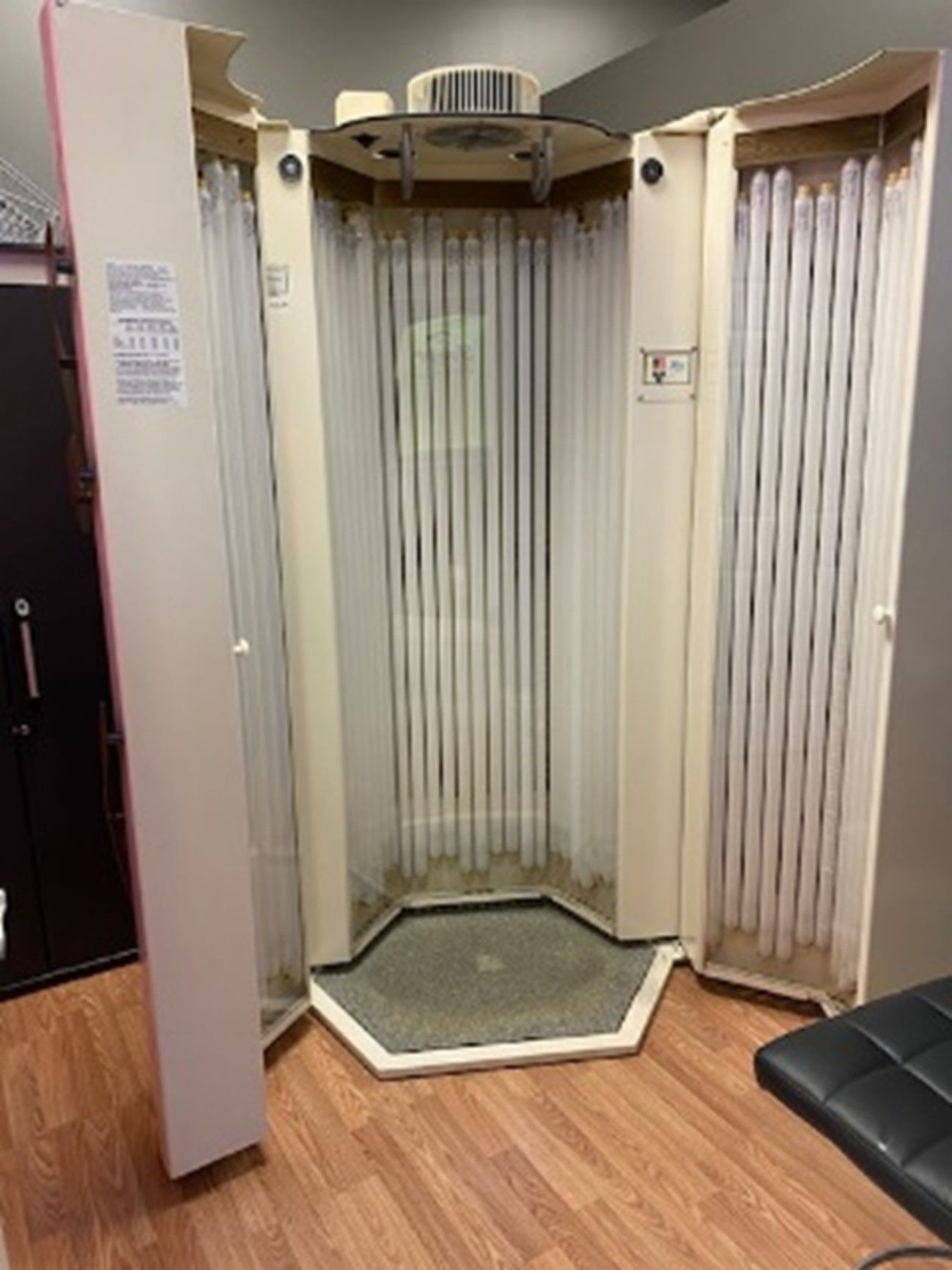 A stand-up tanning bed with open doors, showing numerous UV light tubes inside. It is in a room with wood flooring.