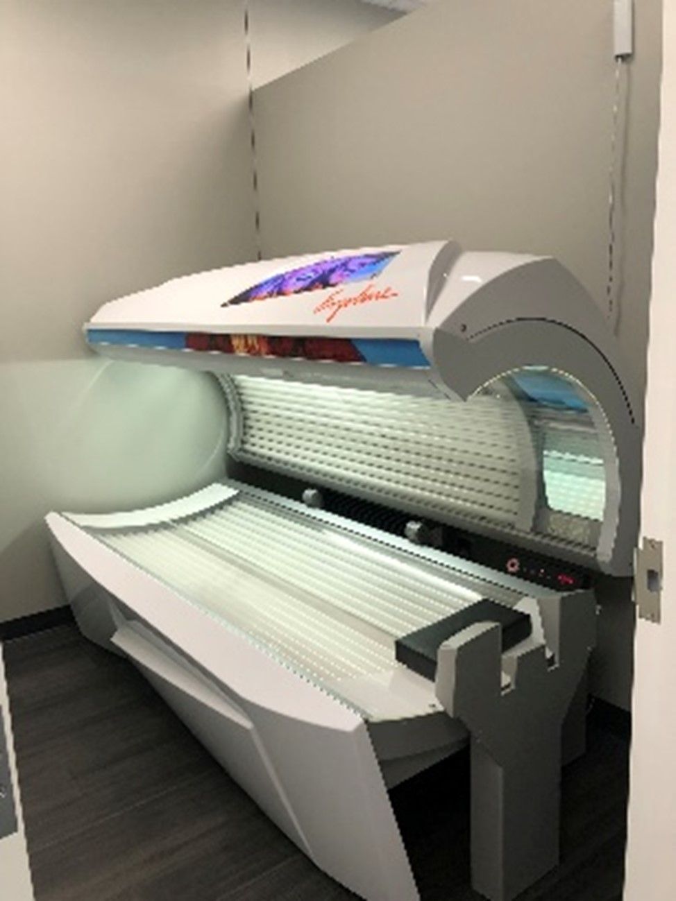 A white tanning bed in a small room with gray floors and white walls; a light panel is visible on the top.