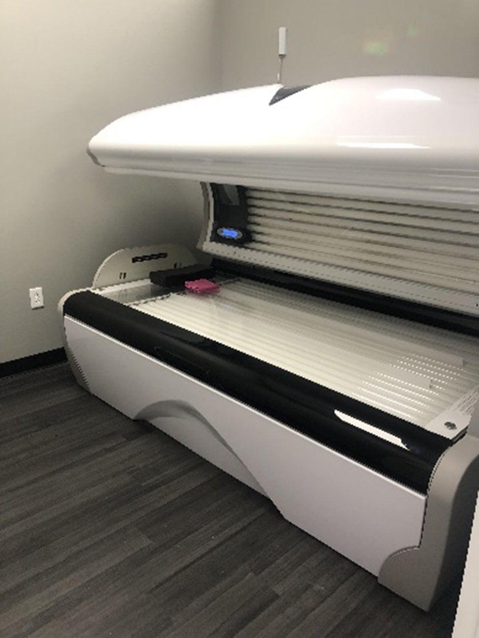 A white tanning bed in a room with gray flooring and walls. The bed is open, revealing the lamps inside.
