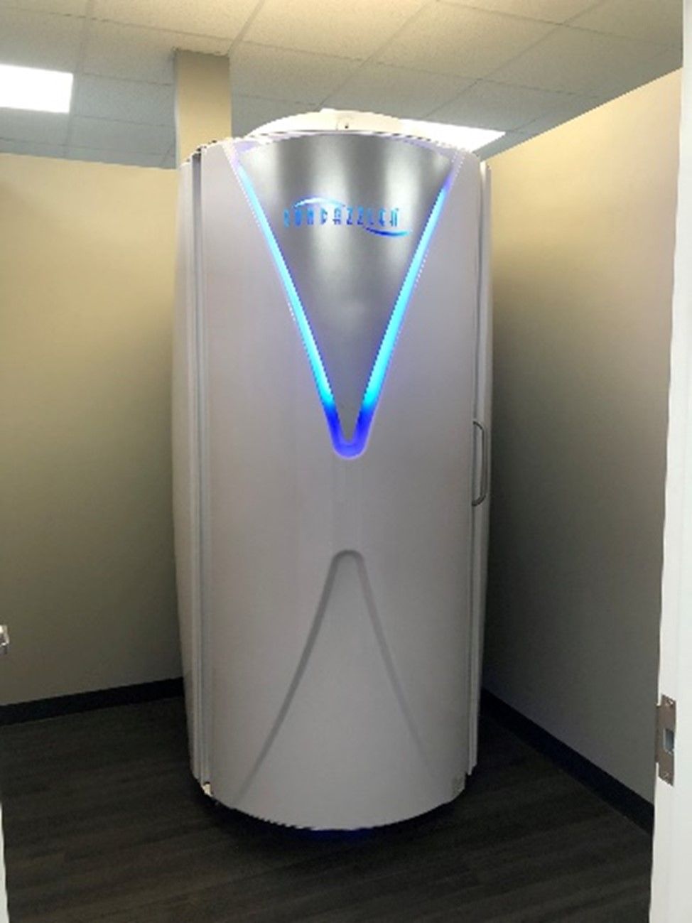 A white stand-up tanning booth with blue lights is situated in a room with neutral-colored walls and a dark floor.