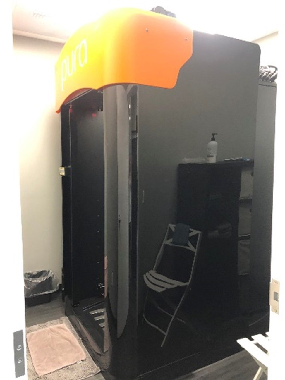 A black spray tan booth with an orange top in a salon. A chair and product are visible.