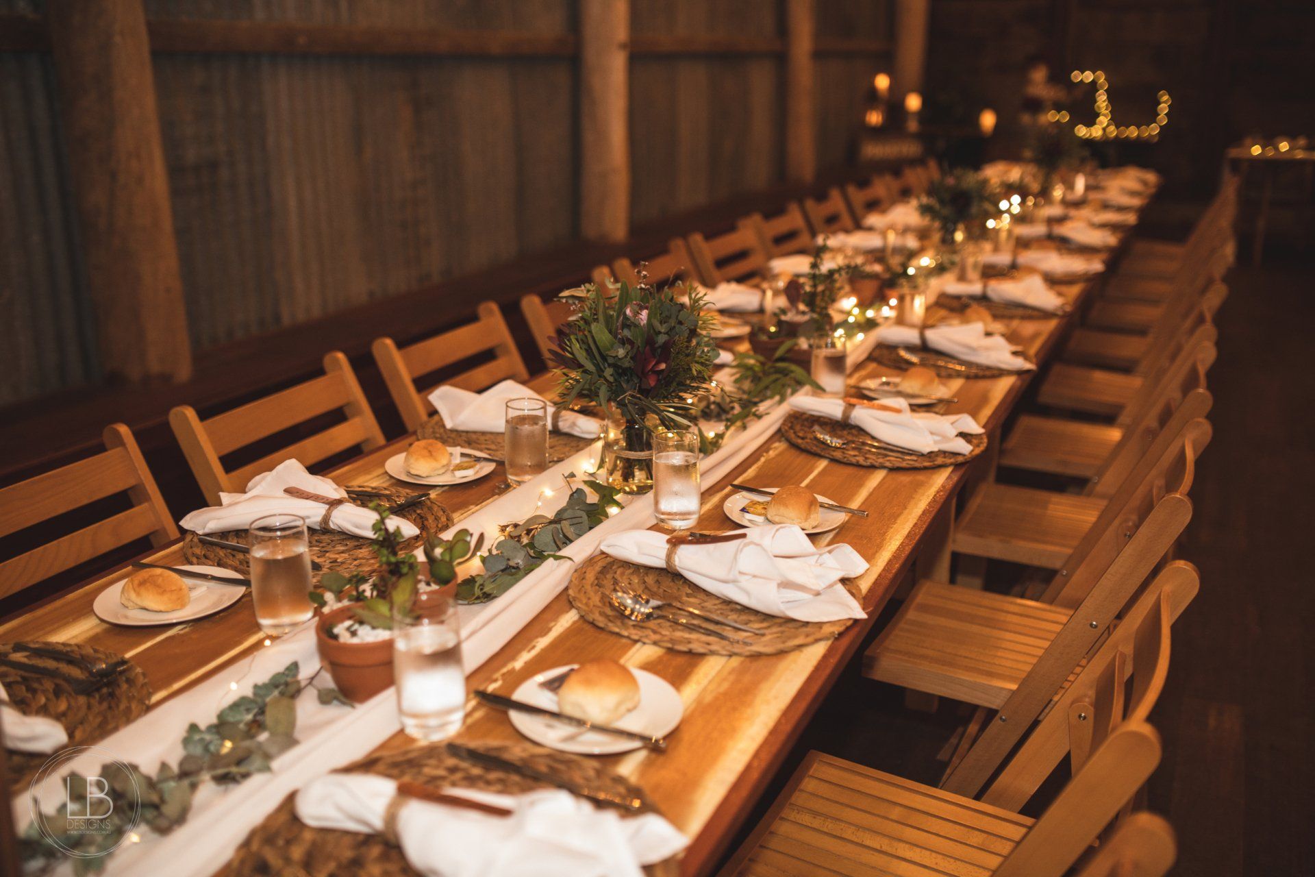 A long wooden table with plates , glasses , and napkins set for a wedding reception.