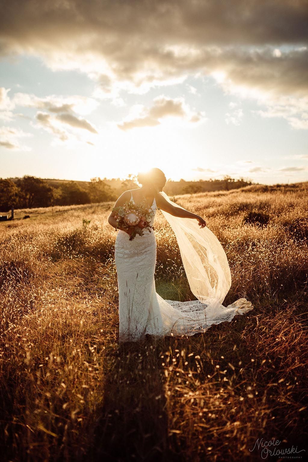 A bride in a wedding dress is standing in a field at sunset.