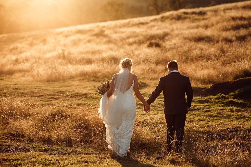 A bride and groom are walking through a field holding hands.