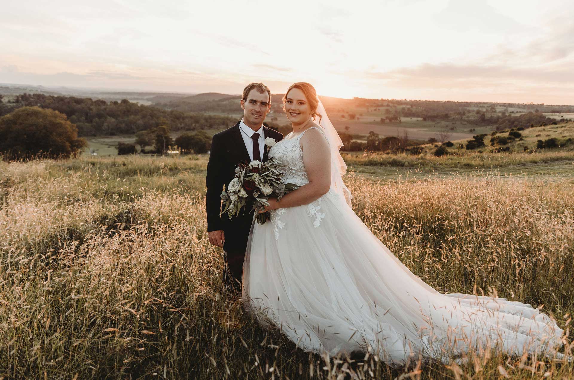 A bride and groom are standing in a field at sunset.
