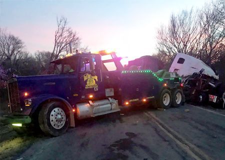 Tow truck at accident scene, night. Truck pulling damaged semi-truck, flashing lights on. Roadside setting, trees in background.