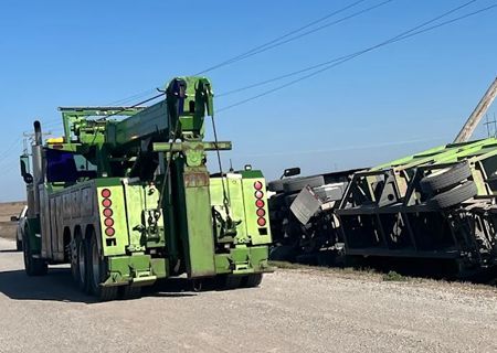 Green tow truck attempting to upright a tipped-over green trailer on a rural road.