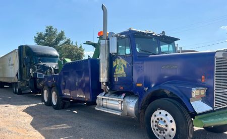 Blue tow truck towing a black semi-truck. Both vehicles are outdoors, under a clear sky.