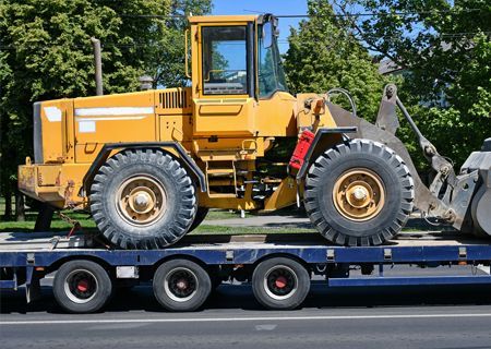 Yellow construction loader on a flatbed trailer, ready for transport on a road with trees.